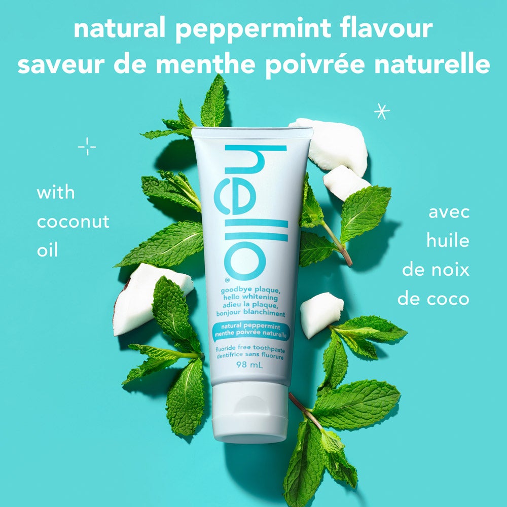 A tube of goodbye plaque hello whitening fluoride free toothpaste stands upright surrounded by fresh mint leaves and coconut pieces on a turquoise background. Text describes "natural peppermint flavour" in English and French, and "with coconut oil" in both languages.